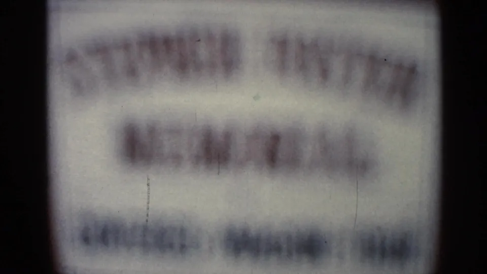 Soft 1966 8mm frame of a sign filling the view, with multiple unreadable text lines and visible film scratches.