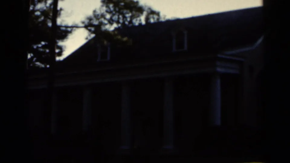 Shadowed 1966 Florida exterior view of a white-columned house framed by trees, with the facade still underexposed.