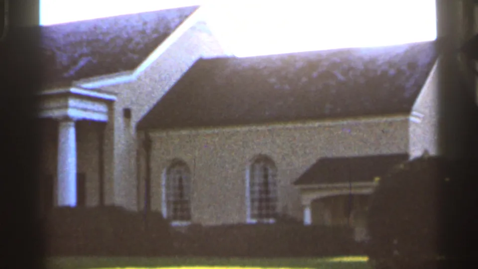 1966 Florida 8mm frame of a stately white building facade with columns, arched windows, shrubs, and bright sky.