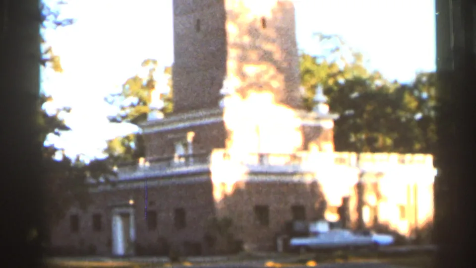 1966 Florida 8mm frame showing a brick building with a prominent tower, white railing, doorway, trees, and sunlit grounds.