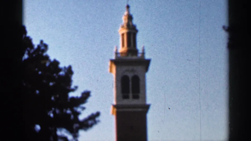 1966 Florida home-movie frame showing an ornate tower viewed from below, framed by dark tree branches and blue sky.