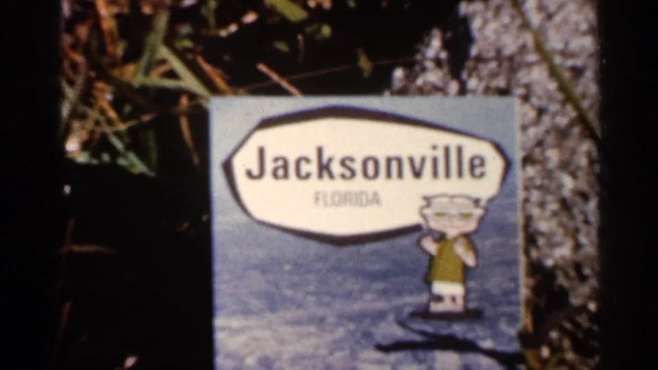 1966 Florida frame showing a Jacksonville, Florida sign with a cartoon mascot holding a sign, surrounded by foliage and tree bark.