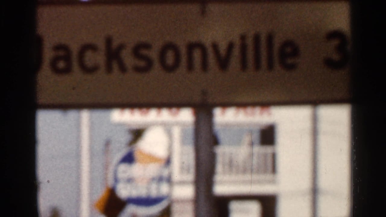 A soft 1960s Florida frame shows a blurred Jacksonville directional sign, with a building facade and another indistinct sign behind it.