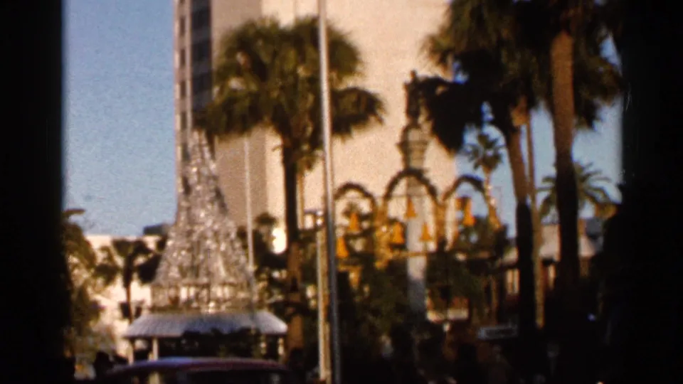The final frame keeps the decorative tree at left with palms, a flagpole, arched public decoration, and indistinct passersby in a Florida street setting.