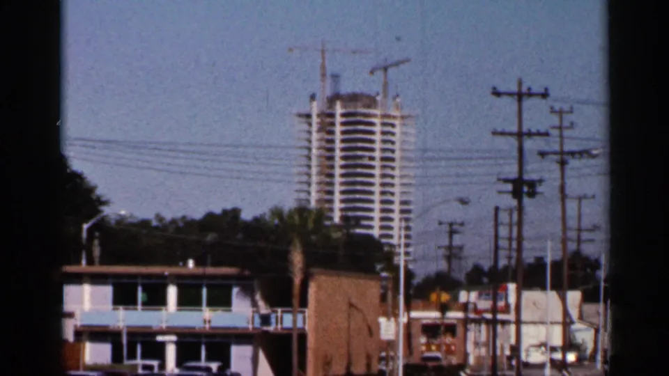 1966 Florida street view showing a tall building under construction, cranes, utility poles, wires, and low roadside structures.