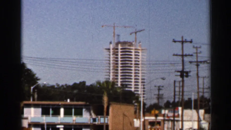 Florida 8mm frame with a high-rise under construction behind roadside buildings, utility poles, wires, and a palm tree.