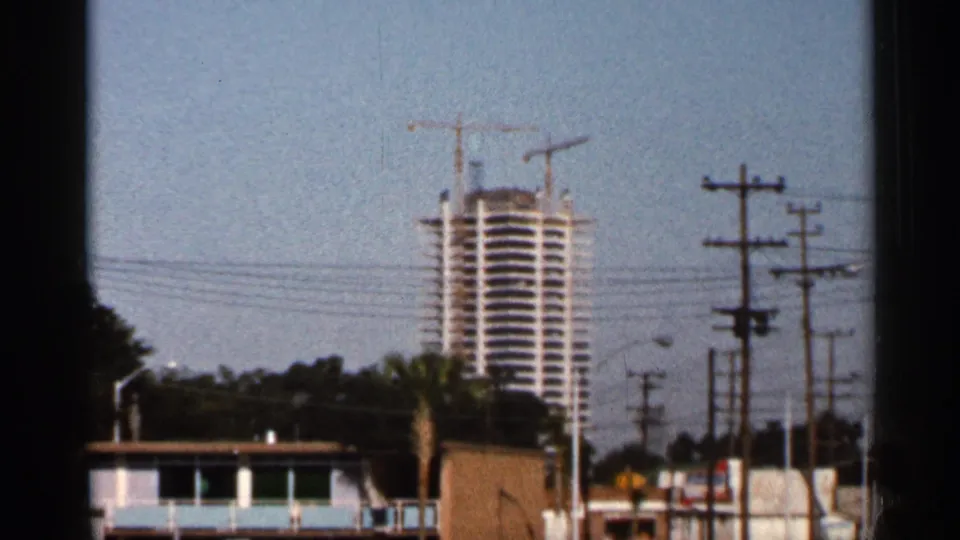 1966 Florida street view with a distant high-rise under construction, cranes, utility poles, palm trees, and roadside structures.