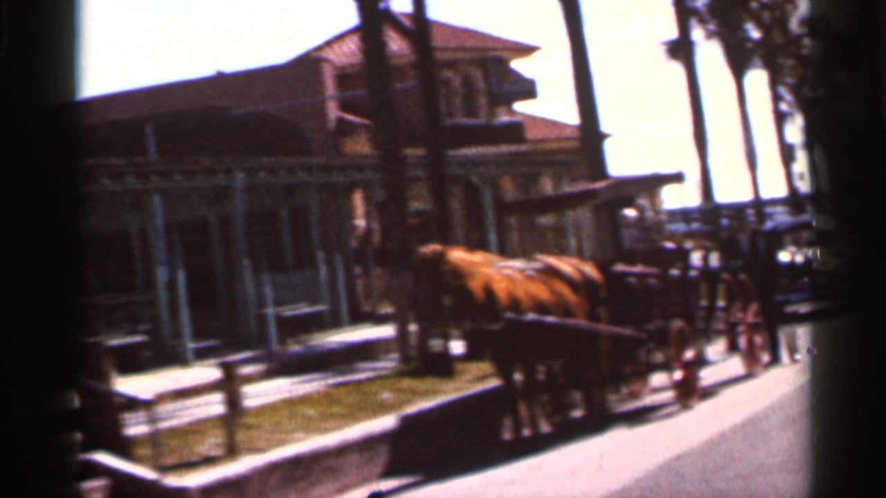 1966 Florida 8mm frame showing a horse-drawn carriage near a building frontage, with a parked car and small figures in the street.