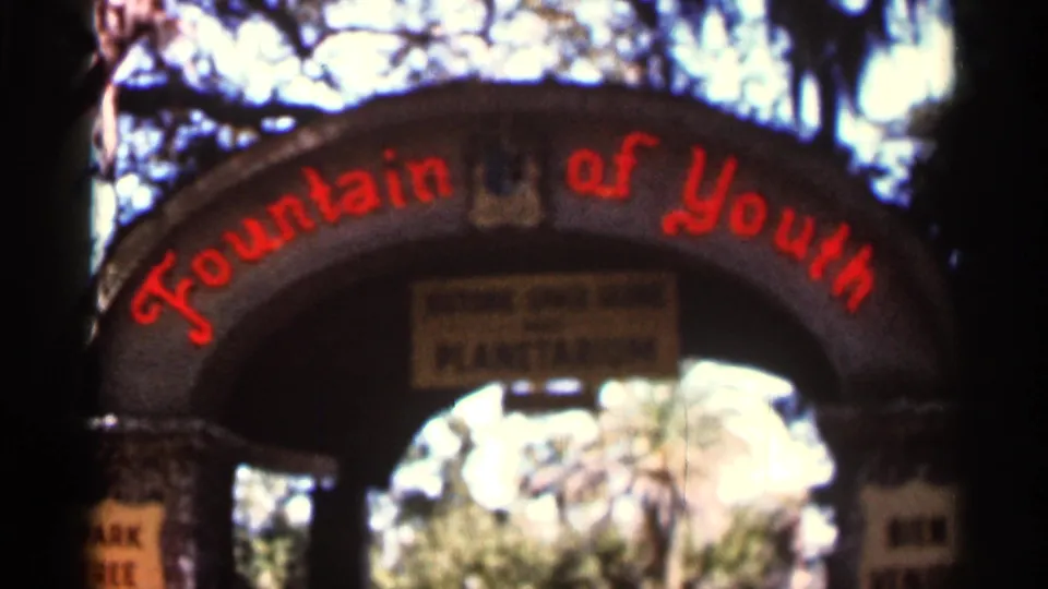 1966 Florida 8mm frame of the Fountain of Youth entrance arch, with red lettering, a hanging sign, trees, and bright sky behind it.