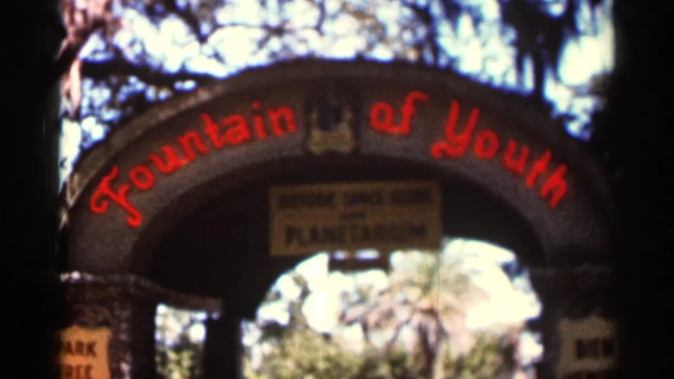1966 Florida 8mm frame showing a closer view of the Fountain of Youth arch, red lettering, shaded columns, hanging sign, and trees beyond.