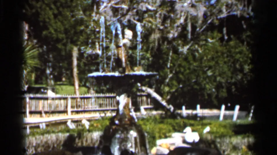 1966 Florida 8mm view of a tiered fountain beside a wooden fence, green lawn, trees, and hanging moss.