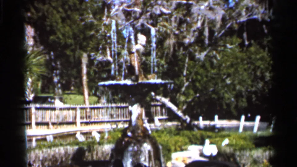 A lush Florida garden scene shows a tiered fountain near fencing and moss-draped trees in 1966 8mm footage.