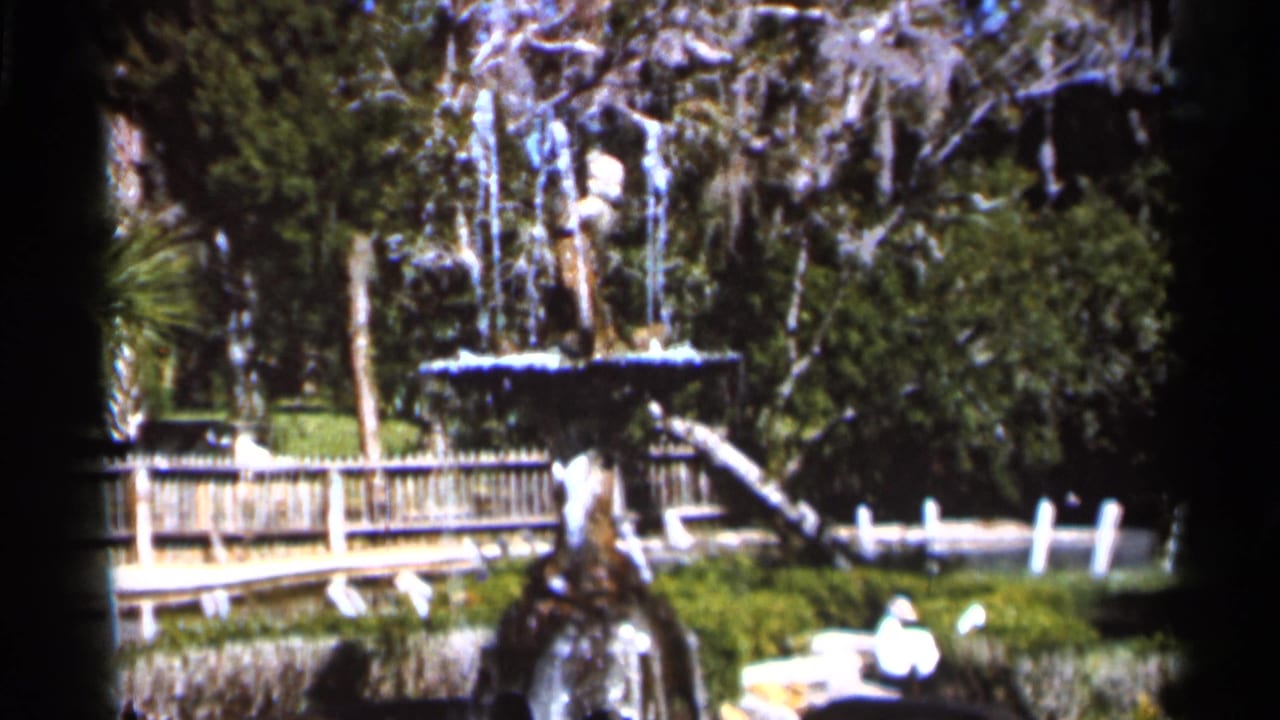 Park fountain with water, wooden railing, greenery, and moss-draped trees.