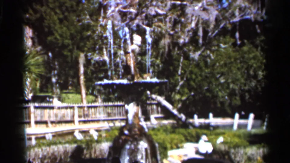1966 Florida 8mm frame showing a sunlit park fountain, wooden railing, shrubs, and trees draped with Spanish moss.
