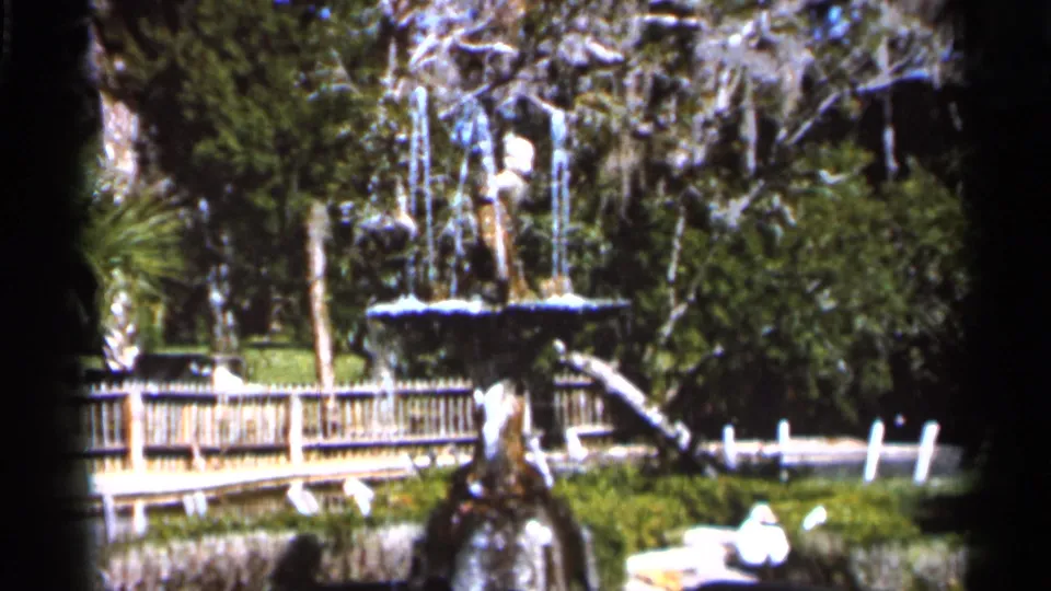 1966 Florida park view with a decorative fountain, rustic wooden railing, dense greenery, and moss hanging from trees.