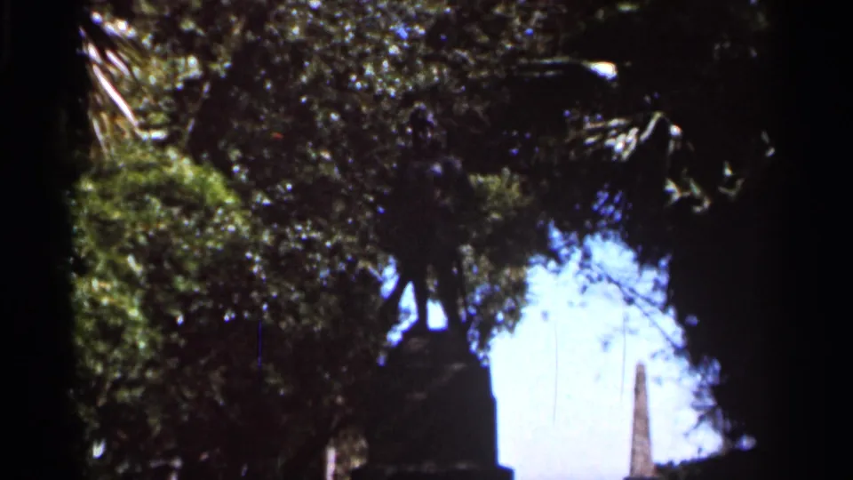 A handheld 1966 Florida view through trees and moss, with a pale sky opening and distant vertical structure or monument.