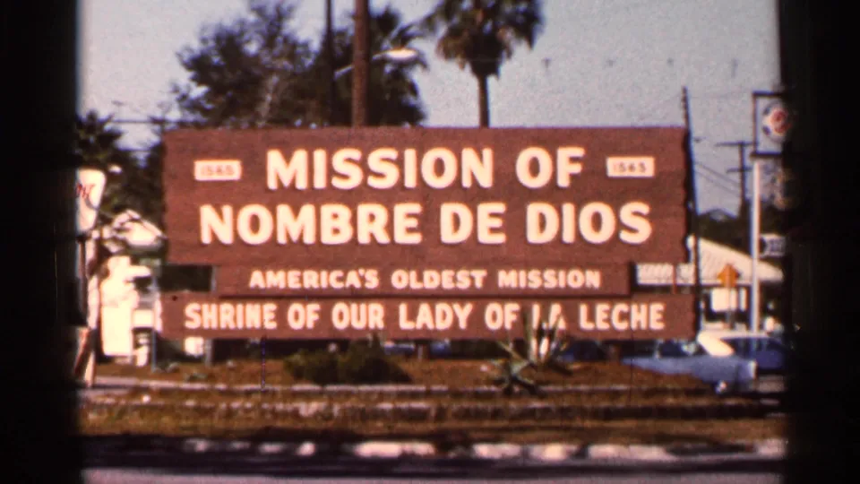1966 Florida roadside sign for Mission of Nombre de Dios, with palm trees and street elements behind it.