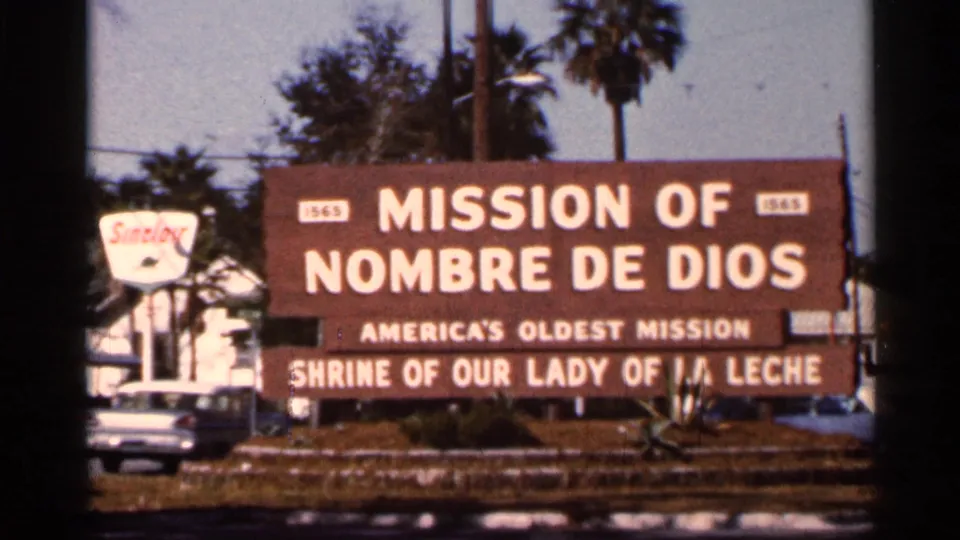 1966 Florida roadside view of the Mission of Nombre de Dios sign, a mid-century car, palms, and a nearby motel-style sign.