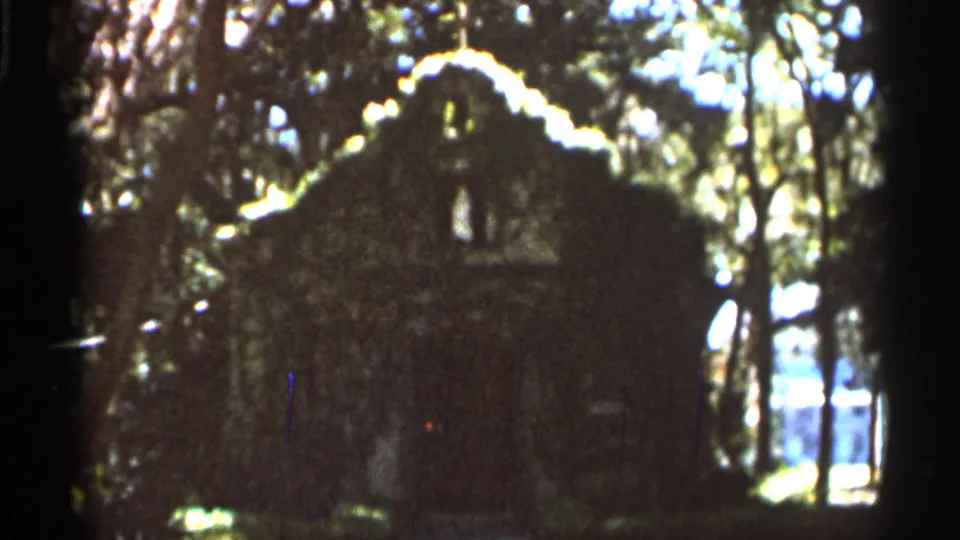 1966 Florida 8mm frame showing a dark cabin-like structure in shaded woods with bright foliage and sky behind it.