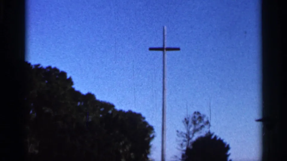 1966 Florida 8mm frame showing a tall wooden cross against a blue sky, with dark trees along the lower edge.
