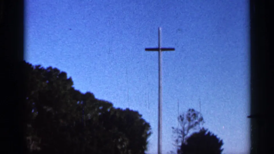 1966 Florida 8mm frame with the wooden cross slightly left of center, blue sky behind it, and trees forming a dark lower border.