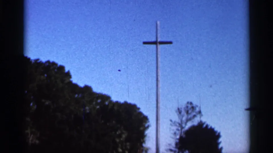 1966 Florida 8mm frame showing the tall cross near center, a blue sky, dark leafy trees, and visible film texture.