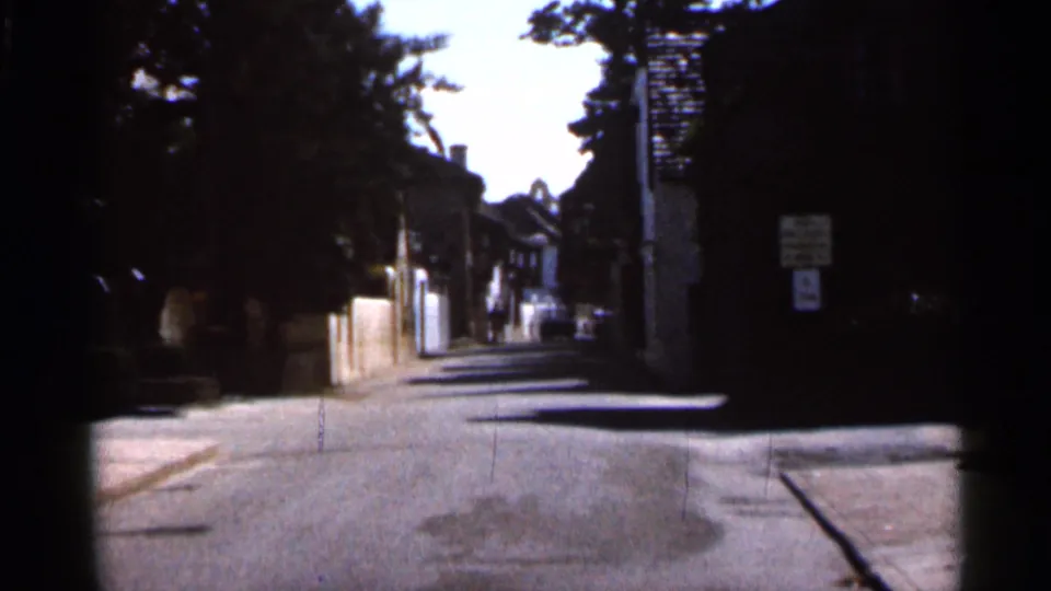 1966 Florida 8mm view down a shaded residential street or alley with trees, walls, signage, and a distant car.