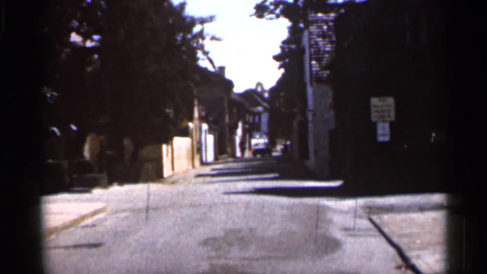 1966 Florida home-movie frame showing a quiet residential street with trees, walls, signs, and a car in the distance.
