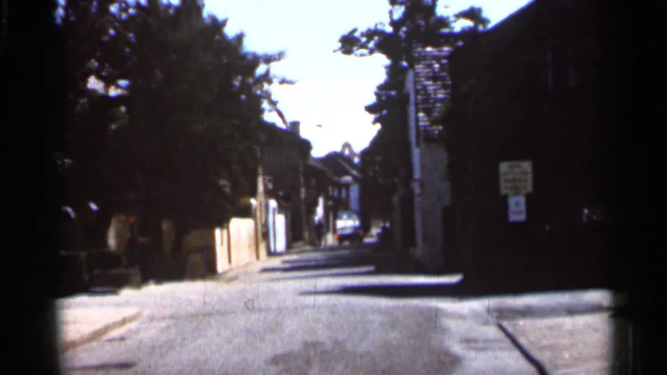 1966 Florida 8mm street view with heavy shadow, low walls, house fronts, signs, and a distant parked car.