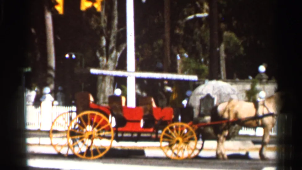 1966 Florida frame of a horse-drawn red buggy with yellow wheels near a white fence and tall cross.