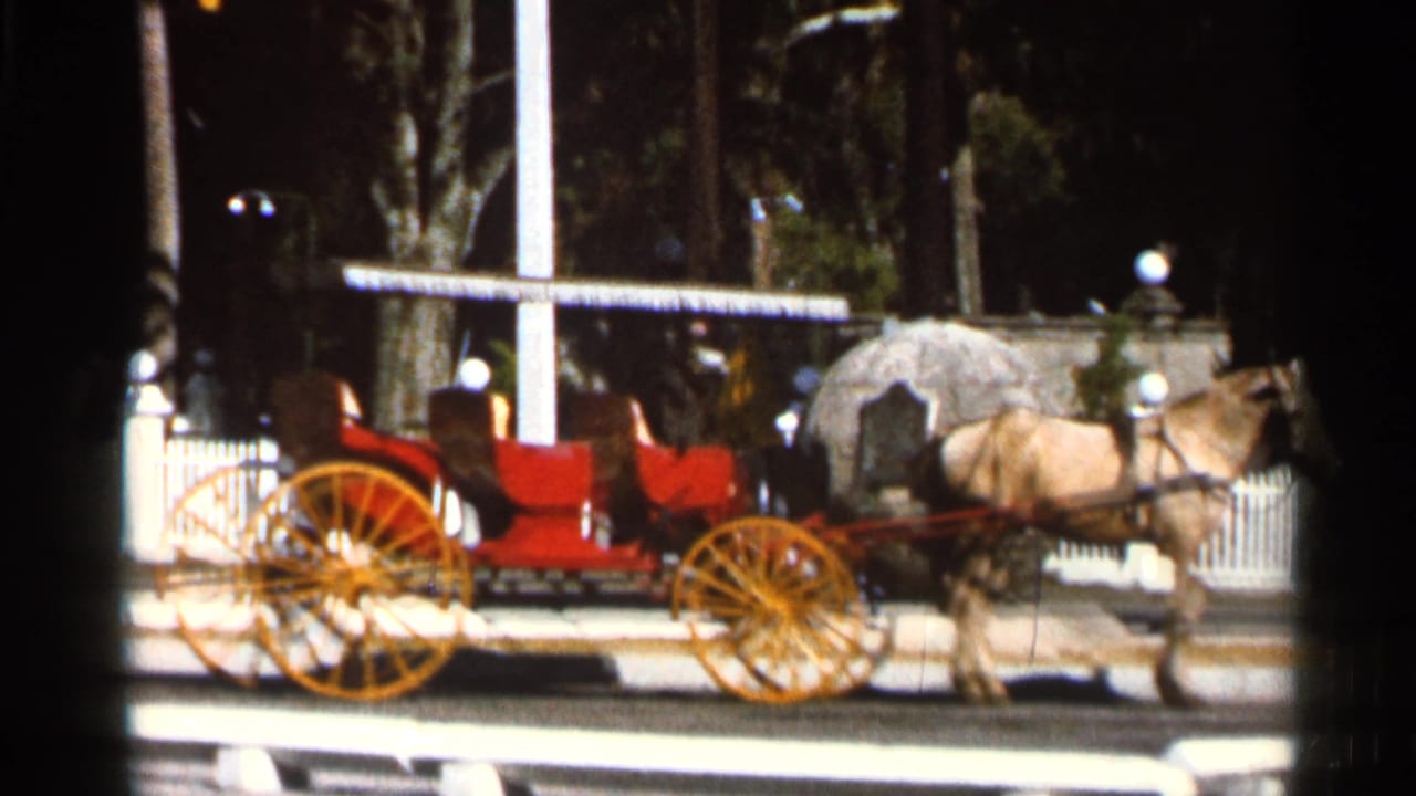 Horse-drawn carriage with red seats and yellow wheels on a street beside a white fence.