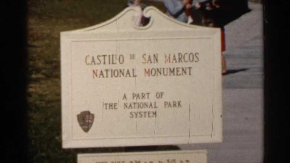 1966 Florida frame showing the Castillo de San Marcos National Monument sign, with partial visitors at the edge of the shot.