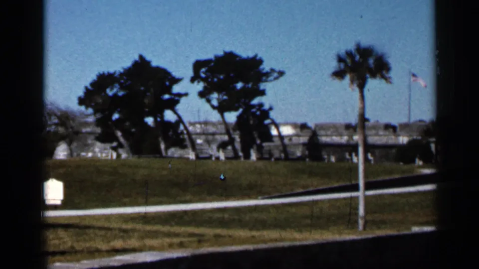 1966 St. Augustine view of Castillo de San Marcos grounds with lawn, trees, fort walls, and an American flag.