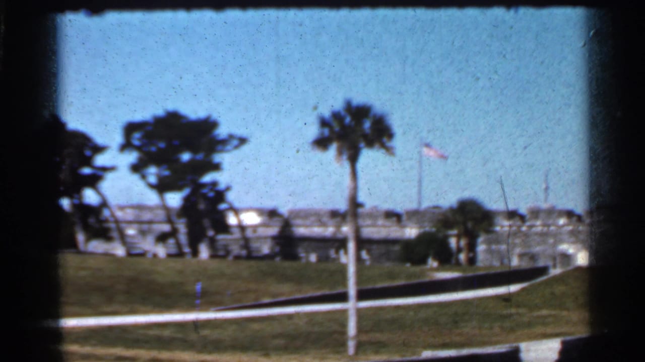 1966 Florida 8mm frame showing a stone military fort beyond a grassy lawn, with palm trees, flagpoles, and a curving approach path.