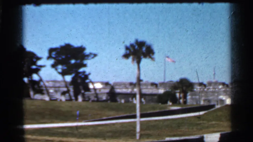 1966 Florida 8mm frame showing a stone military fort beyond a grassy lawn, with palm trees, flagpoles, and a curving approach path.