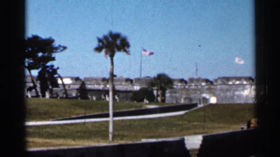 1966 Florida home-movie frame of a stone fort wall with palm trees, a raised lawn, flagpoles, and a bright sky.