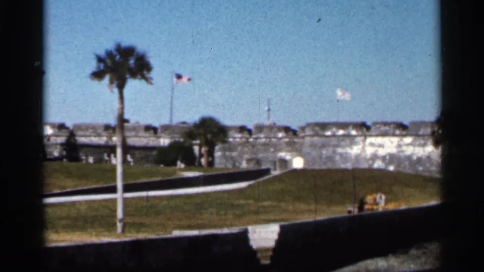 1966 Florida 8mm view of a historic fort area with palm trees, flags, a grassy approach, and a small distant person near the lower frame.