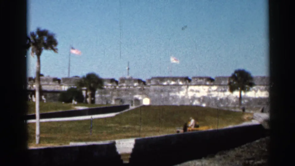 1966 Florida home-movie frame showing the fort wall, palm trees, flying flags, grassy grounds, and small distant visitors near the path.