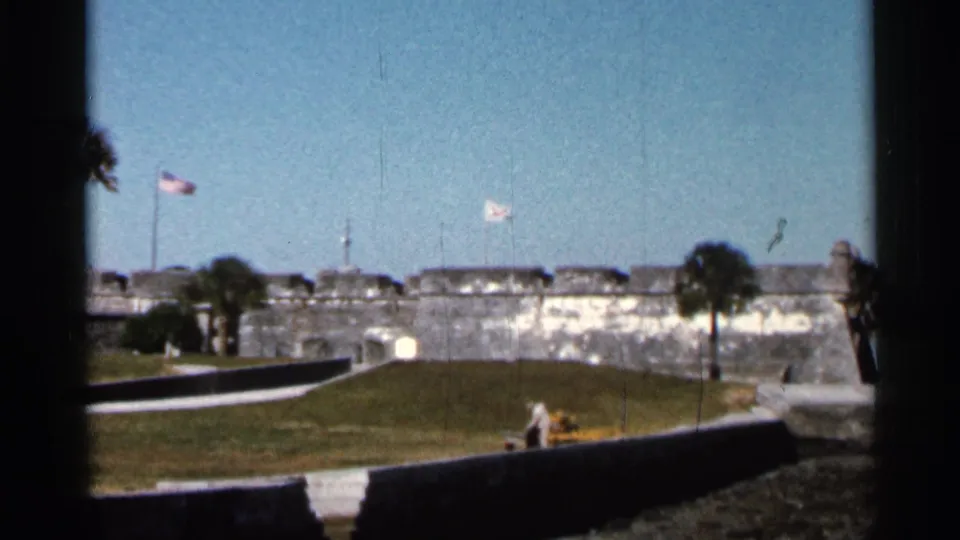 1966 Florida 8mm frame showing a stone fortress, likely Castillo de San Marcos, seen from a visitor viewpoint with flags overhead.