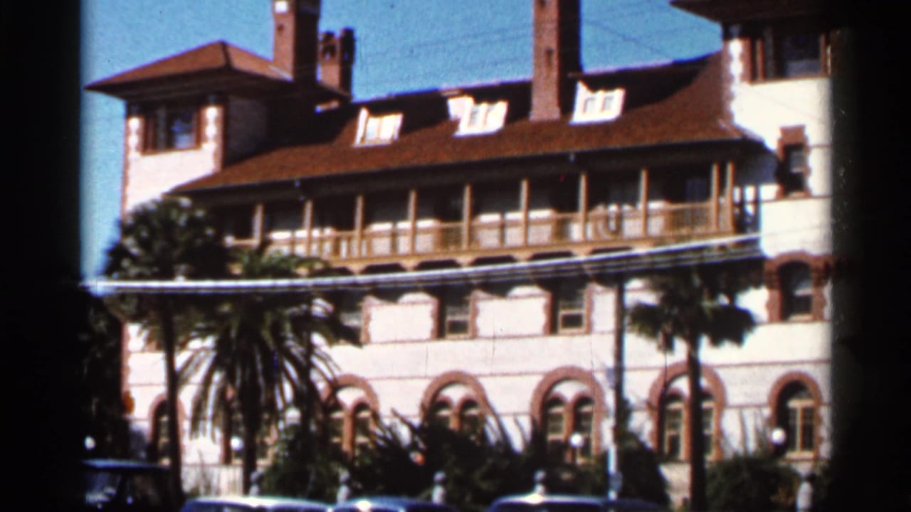 White stucco building with red tile roof, balcony, arched windows, palm trees, and parked cars.