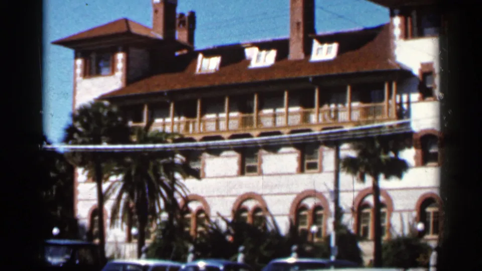 1966 Florida frame of a sunlit white stucco building with red roof, balcony, arched windows, palms, and parked cars at street level.