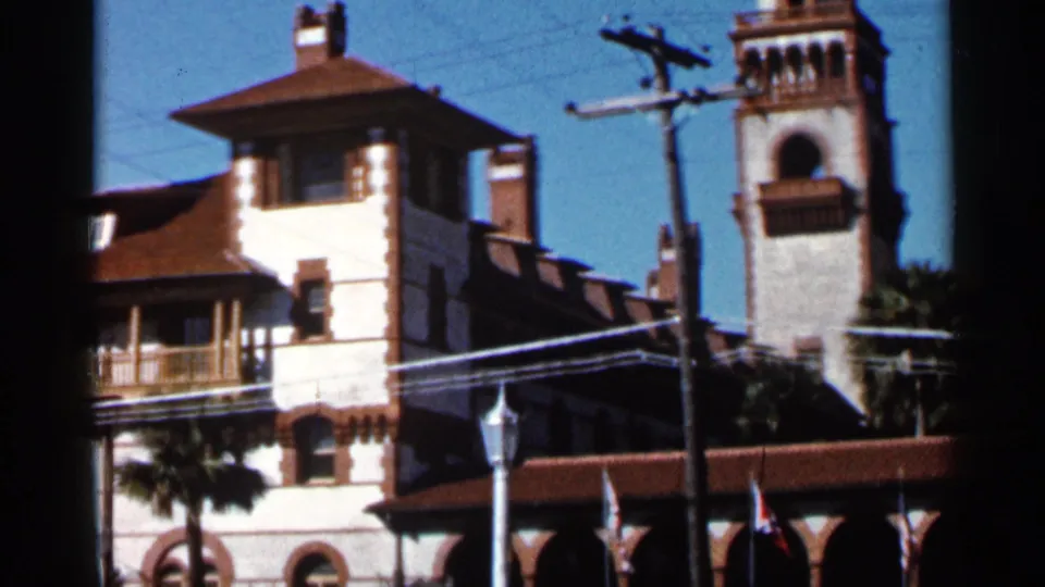 1966 Florida 8mm frame showing the building corner, tower, red tile roofs, overhead wires, street lamp, flags, and palms.