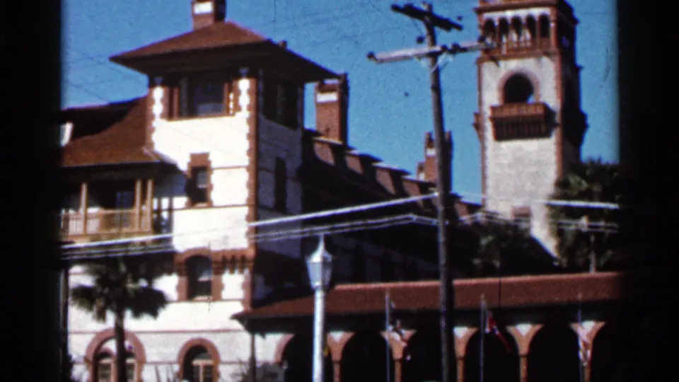 1966 Florida street-level frame with a white stucco building, adjacent tower, red tile roofs, utility lines, street lamp, flags, and palm trees.