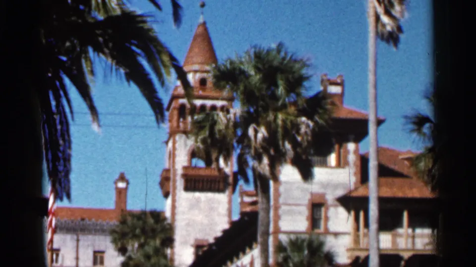 1966 Florida 8mm frame showing the Ponce de Leon Hotel tower, red-tiled rooflines, palms, flagpole, and bright blue sky.