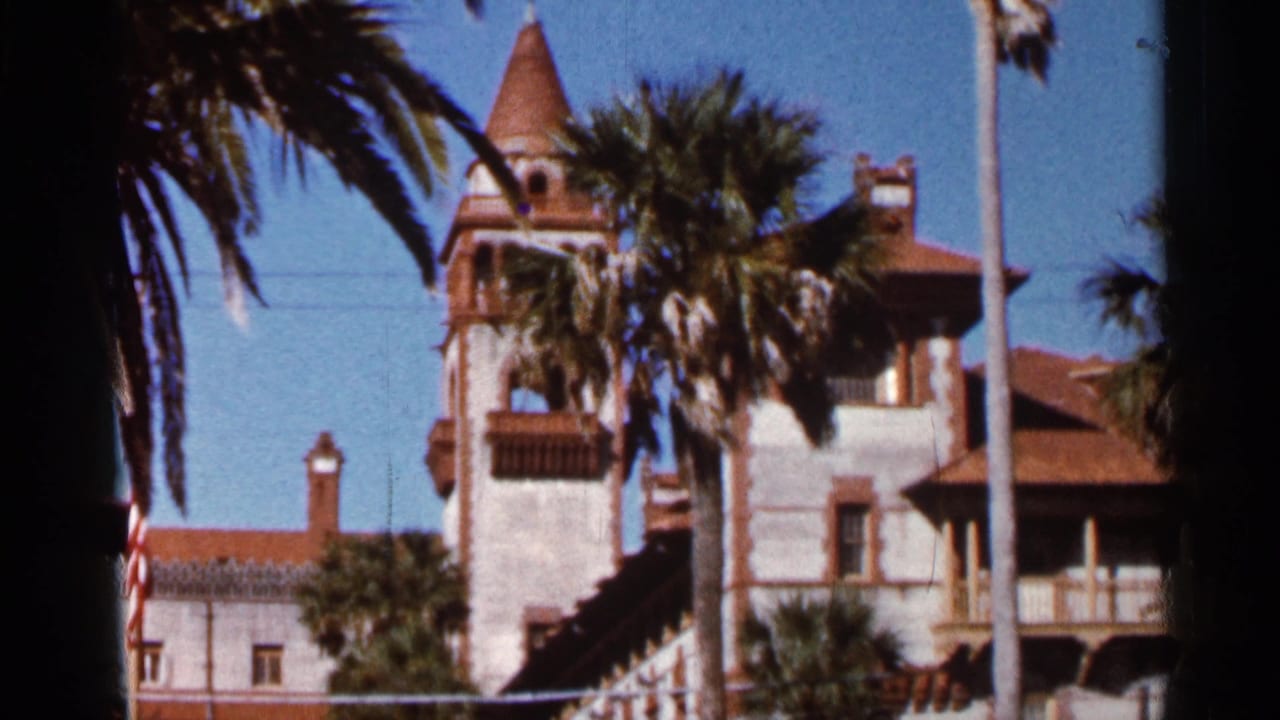 Palm trees frame a light-colored coastal building with red tile roofs under a blue sky.