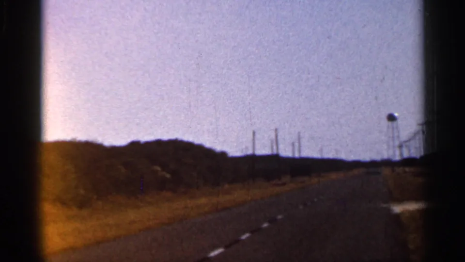 1966 Florida road-trip view from a vehicle, with a two-lane road, dry roadside grass, low vegetation, and distant poles.