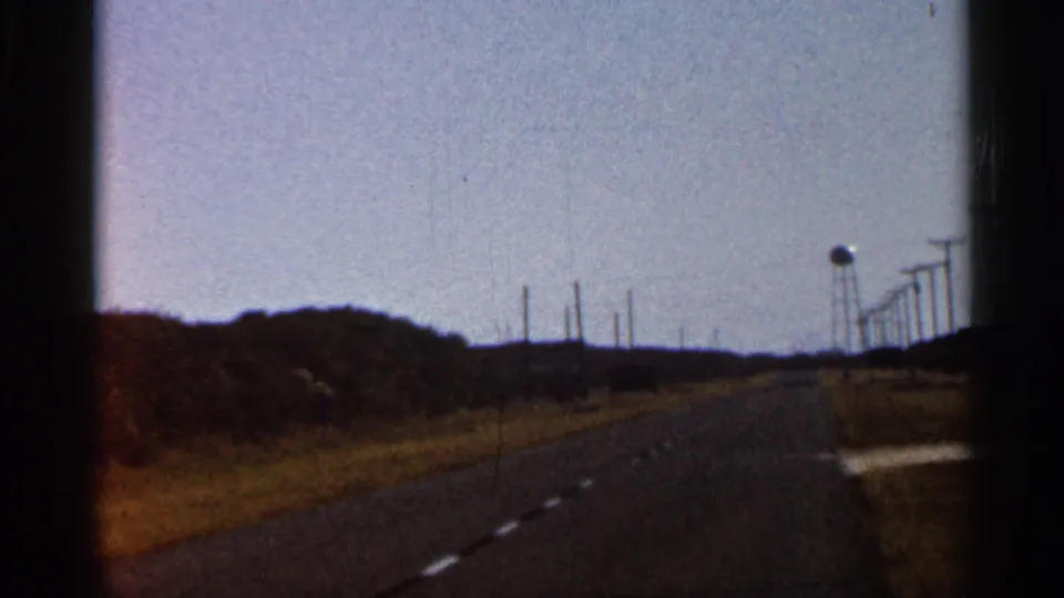 Final 1966 Florida road-trip frame with the road stretching ahead, roadside vegetation, utility poles, and a water tower.