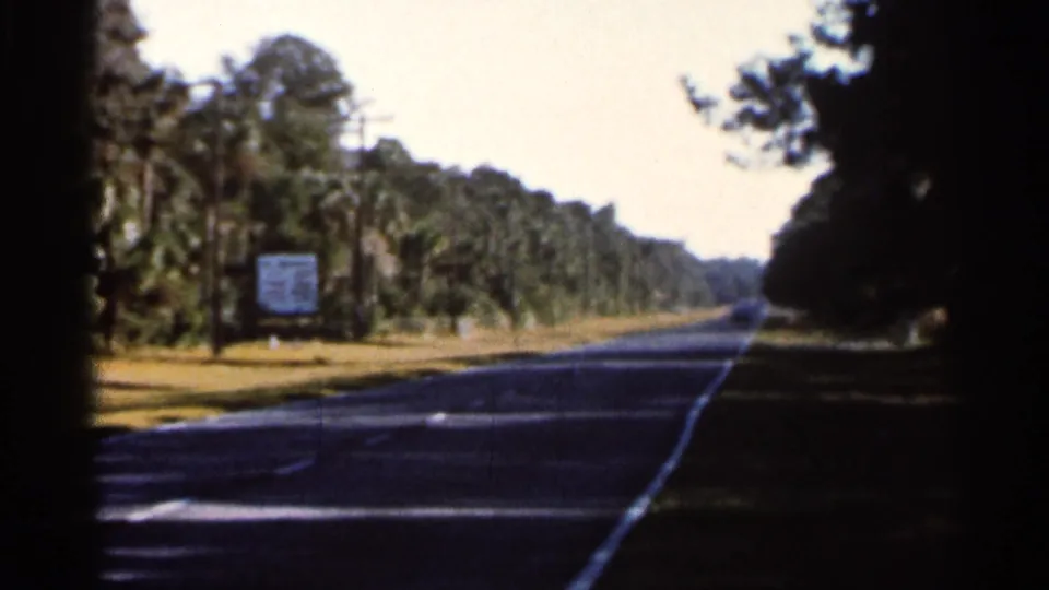 1966 Florida road-trip view from an 8mm home movie, looking down a paved road bordered by trees, palms, and a small roadside sign.