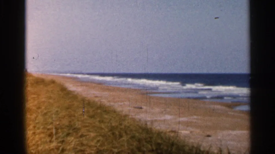 1966 Florida beach view on 8mm film, with golden dune grass in the foreground, pale sand, low surf, open water, and a hazy horizon.