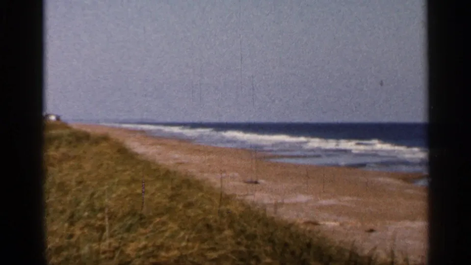 1966 8mm view of a Florida shoreline with grassy dunes, pale sand, ocean surf, and visible film gate edges.